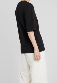 Černé tričko s krátkým rukávem vyrobené z měkkého materiálu s volným střihem, s bočními rozparky pro snadný pohyb. Nosené s bílými kalhotami s širokými nohavicemi.