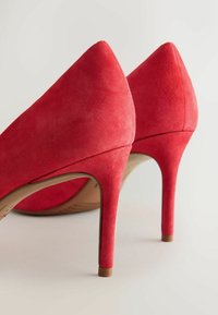 Paire d’escarpins rouges en daim avec bouts pointus et talons fins sur un fond clair et neutre.