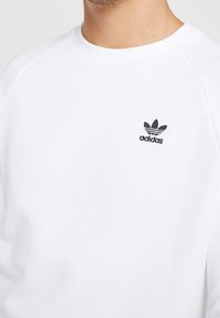 Sudadera blanca con mangas raglán, que presenta un pequeño logo negro de Adidas en la parte izquierda del pecho, hecha de una tela suave.