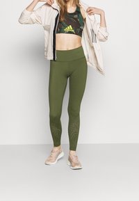 Zielone legginsy z teksturowanymi akcentami, w połączeniu z biustonoszem sportowym w kamuflaż, który ma jasne logo, oraz lekką kurtką. Różowe sneakersy dopełniają całość.