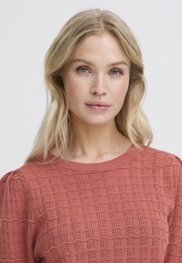 Blonde Frau mit blauen Augen, die einen strukturierten korallenfarbenen Strickpullover trägt und vor einem schlichten hellgrauen Hintergrund nach vorne schaut.