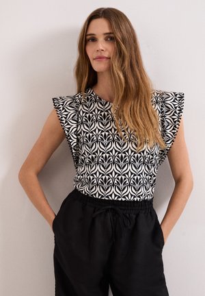 Woman with long hair wearing a black and white patterned sleeveless top and black drawstring pants, standing against a plain wall.