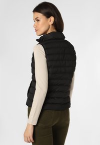 Czarna kamizelka puchowa z wysokim kołnierzem, występująca w pikowanym wzorze i slim fit. Nosi na sobie jasnoskalowy T-shirt z długim rękawem oraz oliwkowe spodnie.