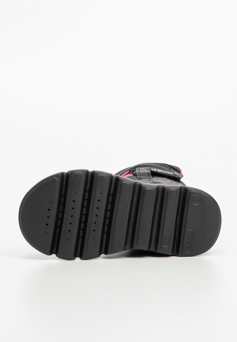 Suola di sneakers nera con solchi profondi e piccole perforazioni, inclinata verso l'alto, che mostra parte della tomaia con dettagli neri e rosa.