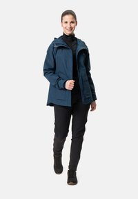 Giacca impermeabile blu scuro con cappuccio, zip frontale e tasche laterali. Indossata sopra una maglietta nera e abbinata a pantaloni e scarpe neri.