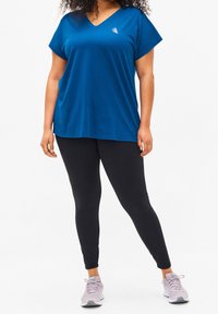 Niebieski t-shirt z dekoltem w serek i krótkimi, podwiniętymi rękawami, zestawiony z czarnymi legginsami. Koszulka ma małe logo na piersi i luźny krój.