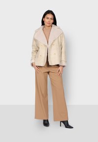 Giacca beige doppiopetto con colletto di pelliccia color crema, abbinata a un maglione a coste color marrone chiaro e pantaloni a gamba larga color marrone chiaro. Stivaletti neri con tacco.