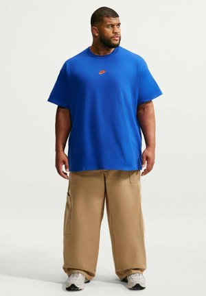 Uomo in piedi che indossa una maglietta Nike blu, pantaloni cargo beige larghi e scarpe da ginnastica bianche, guarda verso destra su uno sfondo semplice.