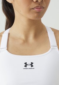 Hvit sports-bh med justerbare hvite stropper. Har en svart Under Armour-logo i midten. Glatt tekstur, tettsittende design.
