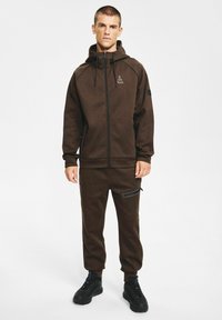 Brązowa bluza na zamek z kapturem i sznurkami, dopasowane brązowe spodnie dresowe oraz czarne akcenty. Noszona z czarnymi butami sportowymi, o smukłym kroju.