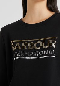 Černá mikina s žebrovaným výstřihem a výrazným textem ve zlaté a stříbrné barvě. Text obsahuje nápisy "BARBOUR" a "INTERNATIONAL" ve stylizovaných fontech.