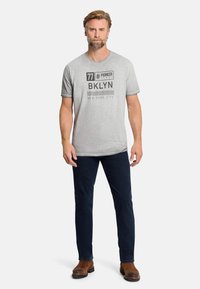 Graues Baumwoll-T-Shirt mit einem grafischen Druck, der "77 PIONEER BKLYN NEW YORK CITY" zeigt. Kombiniert mit dunkelblauen Jeans und braunen Stiefeln.