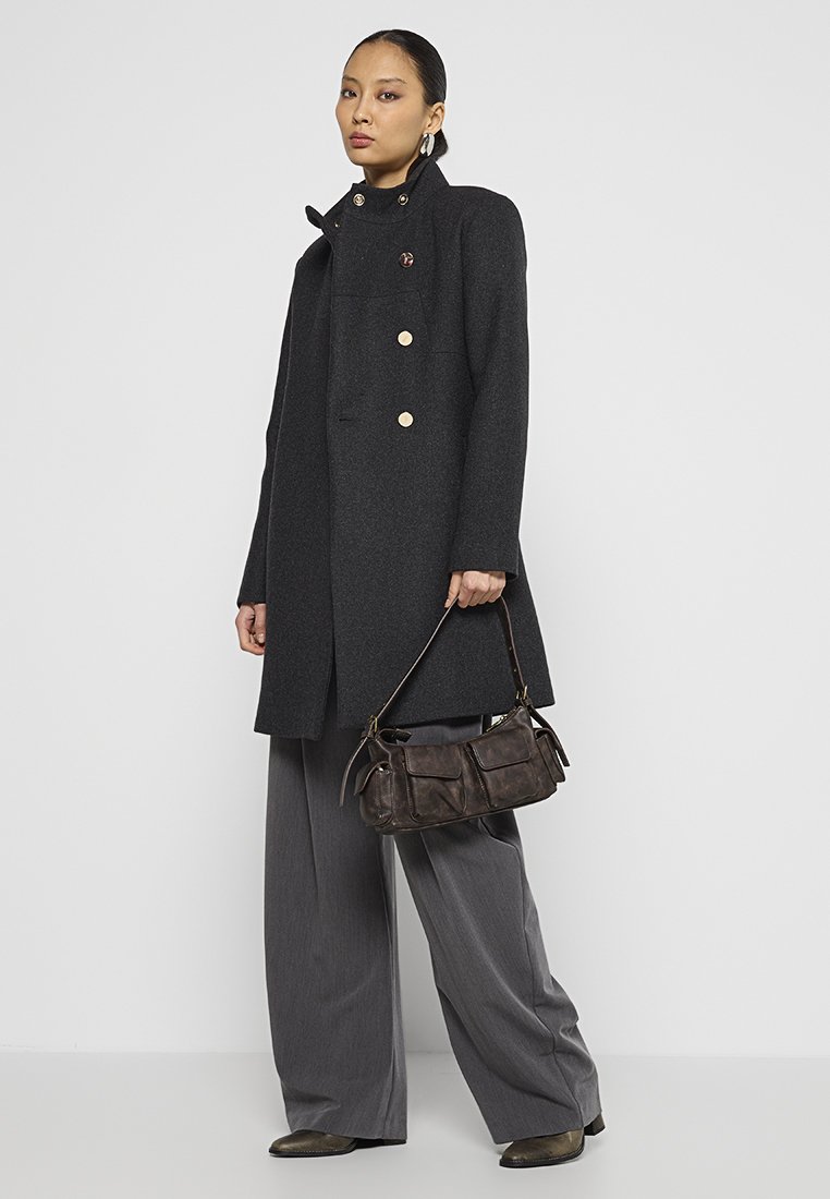 Cappotto di lana nero con colletto alto, bottoni dorati e una texture liscia. Abbinato a pantaloni grigi a gamba larga e una borsa in pelle marrone.