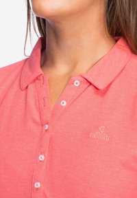 Koraalkleurig poloshirt van textuurstof, met een kraag, een knoopsluiting aan de voorkant en klein geborduurd "Nomad"-logo. De knopen zijn wit.
