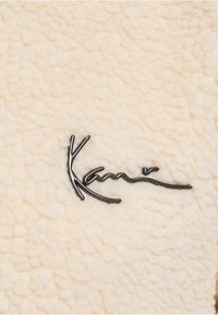 Krämfärgad texturerad tyg med en fluffig yta, prydd med en mörk metallisk logotyp "Kani" i en elegant stil.