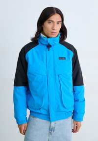 Blaue und schwarze isolierte Jacke mit hohem Kragen, Reißverschluss vorne, zwei Fronttaschen und elastischen Bündchen. Mit Logo auf der Brust.