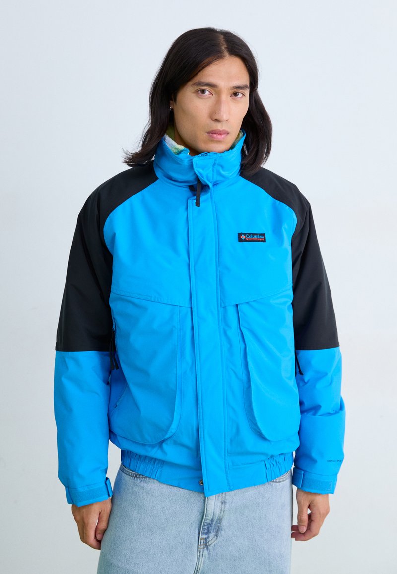 Blaue und schwarze isolierte Jacke mit hohem Kragen, Reißverschluss vorne, zwei Fronttaschen und elastischen Bündchen. Mit Logo auf der Brust.