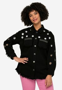 Zwarte denim blouse met zilveren rings en studs, twee voorzakken, franjes aan de onderkant, lange mouwen en knoopsluiting. G gedragen met roze broek.