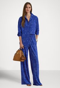 Modrá košeľa s paisley vzorom a široké nohavice z ľahkej tkaniny, doplnené hnedou kabelkou so zlatým kovaním a farebným príveskom.