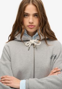 Grauer Reißverschluss-Hoodie mit Kordelzugkapuze, der eine weiche Textur aufweist, ein gesticktes Logo auf der Brust hat und mit einem hellblauen Hemd darunter kombiniert ist.