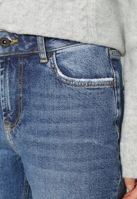Jeans de ganga em um desbotado azul claro com corte reto. As características incluem um bolso frontal, costura dourada e um fecho de botão metálico.