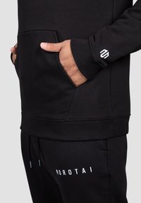 Hanorac negru cu buzunar frontal, manșete ribbate și detaliu cu logo. Asortat cu pantaloni de trening negri care au inscripții albe ale brandului.