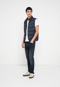 Granatowa pikowana kamizelka puchowa z wysokim kołnierzem i suwakiem z przodu. Nosi się ją na białej koszulce T-shirt, w połączeniu z ciemnymi dżinsami i jasnymi trampkami.