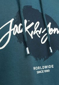 Sudadera verde azulado con texto blanco "Jack & Jones" y "WORLDWIDE SINCE 1990". Presenta cordones y un diseño salpicado en la parte delantera.