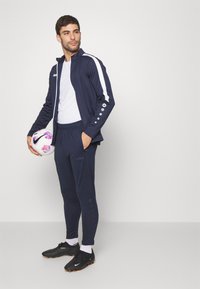 Marineblauwe sportjack met witte strepen, zwarte fitted broek, zwarte voetbalschoenen, met een gemusterde voetbals bal in de hand. Bevat logo-accenten.