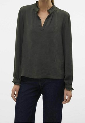 Vrouw draagt een donkergroene blouse met lange mouwen, gerimpelde kraag en manchetten, gecombineerd met donkerblauwe jeans tegen een effen achtergrond.