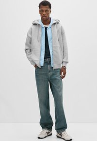 Young man wearing a light gray hoodie over a blue striped shirt, loose blue jeans, white sneakers, and silver wrist accessories against a white background.