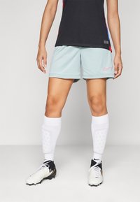 Lichtblauwe Nike short met roze logo, gecombineerd met witte kniekousen en zwart-witte voetbalschoenen. Glad materiaal, atletische pasvorm.