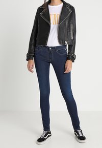 Chaqueta de moto de cuero negra sobre una camiseta blanca con texto amarillo, combinada con jeans ajustados azul oscuro y zapatillas negras.