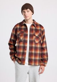 Camicia a quadri in tonalità arancioni, marroni e crema, con due tasche sul petto, chiusura a bottoni e vestibilità rilassata. Tessuto morbido e strutturato.
