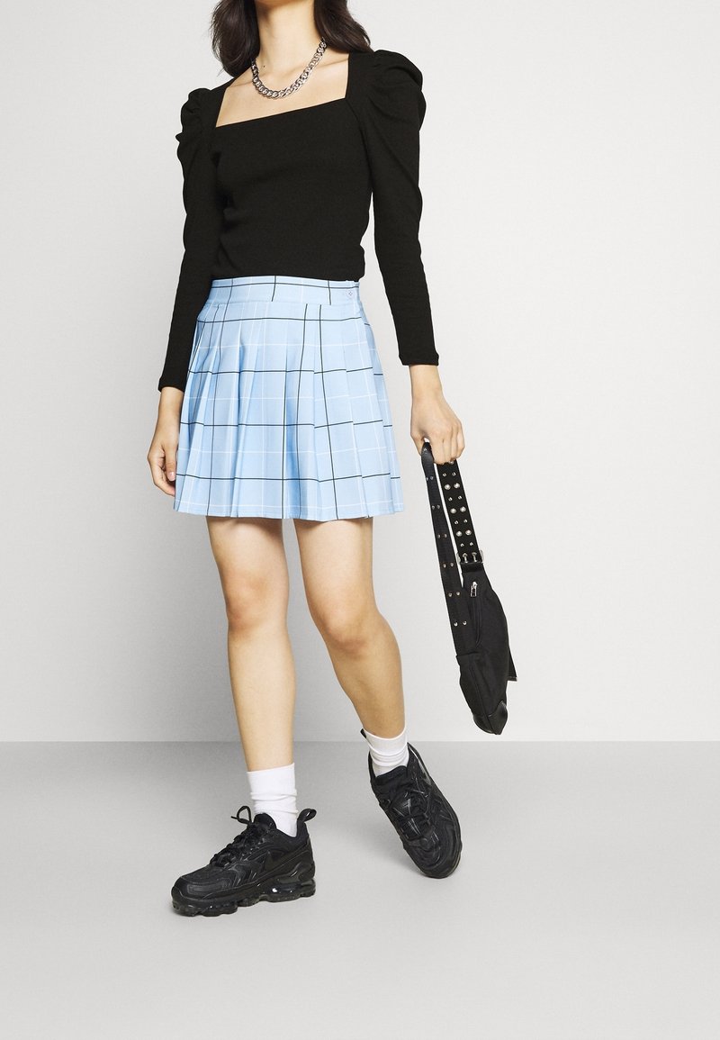 Svart långärmad topp med puffärm, ljust blå rutig plisserad kjol, svarta sneakers och en liten svart väska med nitar.