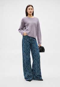 Γυναίκα στέκεται φορώντας μωβ μάλλινο πουλόβερ, φαρδιά μπλε παντελόνια με σχέδια, μαύρα παπούτσια, κρατώντας μια μικρή μαύρη τσάντα, μπροστά σε λευκό φόντο.