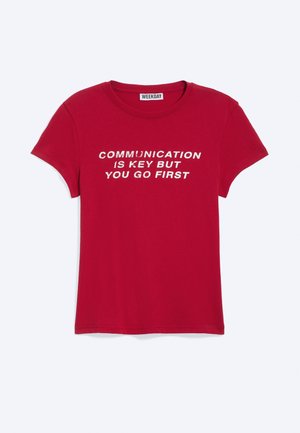 T-shirt rouge à manches courtes avec texte blanc « LA COMMUNICATION EST LA CLÉ MAIS TU Y VAS EN PREMIER » et une étiquette de la marque WEEKDAY au col.