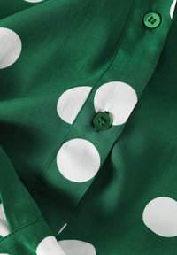 Žalia medžiaga su dideliais baltais taškeliais, puošta žaliais mygtukais. Lygi tekstūra su matomomis siūlėmis ir sulankstytais kraštais.