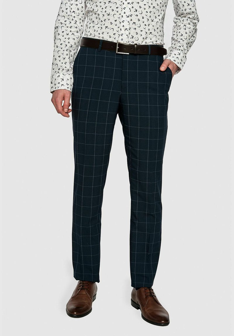 Granatowe spodni w kratę o dopasowanym kroju, wykonane z lekkiego materiału. Zawierają pasek z metalową klamrą w kolorze srebrnym oraz brązowe skórzane buty.