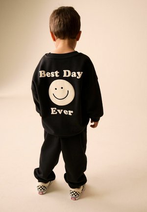 Kind trägt schwarzen Pullover mit "Best Day Ever" und einem Smiley auf dem Rücken, schwarze Hose und karierte Schuhe, steht auf beigem Boden.