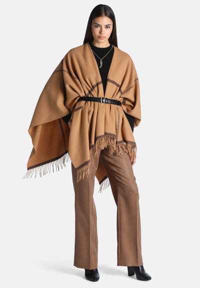 Femme portant un poncho marron à franges avec une ceinture noire, un haut noir, un pantalon marron, des bottes noires et un collier pendentif, debout sur un fond blanc.