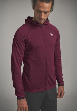 Mann trägt einen bordeauxroten Kapuzenpullover mit Reißverschluss und kleinem weißem Logo auf der Brust, schaut nach unten, die Hände locker an den Seiten.