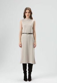 Beige mouwloze jurk van gestructureerde stof, in de taille samengebonden met een donkere riem met een decoratieve gesp, gecombineerd met donkere kniehoge laarzen.