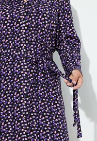 Paarse button-up jurk met een zwart en lavendel stippenpatroon, vastgebonden in de taille met een bijpassende riem. Gladde stoftextuur.