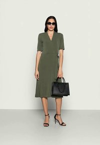 Vestido envolvente verde oliva com mangas curtas, cintura ajustada e comprimento até ao joelho; combinado com uma bolsa preta e sandálias pretas com tiras.