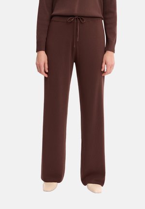 Osoba nosząca brązowe spodnie o szerokich nogawkach z troczkiem w talii i pasującą brązową bluzkę, do tego jasnobeżowe buty, stojąca na białym tle.