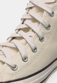 Κοντινό πλάνο σε πάνινο αθλητικό παπούτσι κρεμ χρώματος με λευκά κορδόνια περασμένα από ασημί ροδέλες και λεπτομέρειες ραφής κατά μήκος της άκρης.