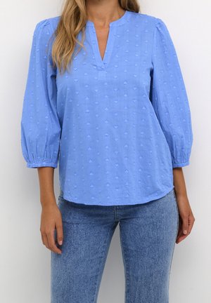 Kvinde iført en lyseblå tekstureret bluse med 3/4-pufærmer og blå jeans mod en hvid baggrund.