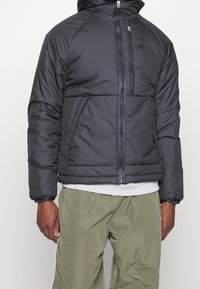 Svart puffjacka med huva, med en framdragkedja, elastiska ärmslut och en bröstlogga. Bärs över en grå skjorta och olivgröna byxor.