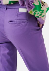 Pantalon violet en tissu lisse, avec une poche arrière fendue. Une étiquette sur la ceinture indique "BRAX FEEL GOOD".